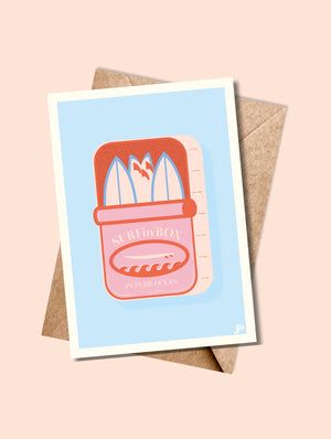 Carte postale SURFin'BOX bleu ciel accompagnée de son enveloppe kraft, posée sur une table estivale.